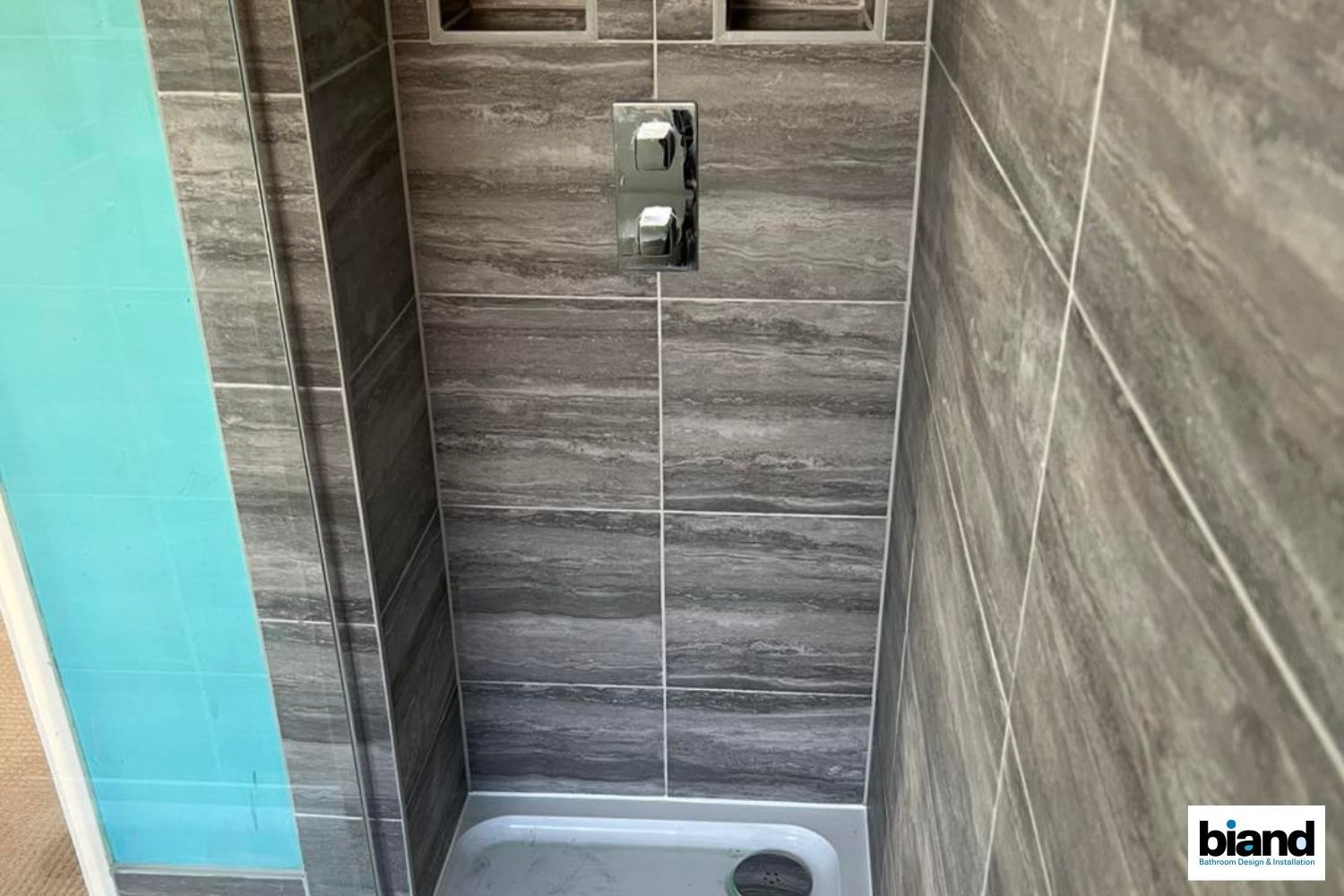 Gray tiled shower with silver fixtures and teal accent.