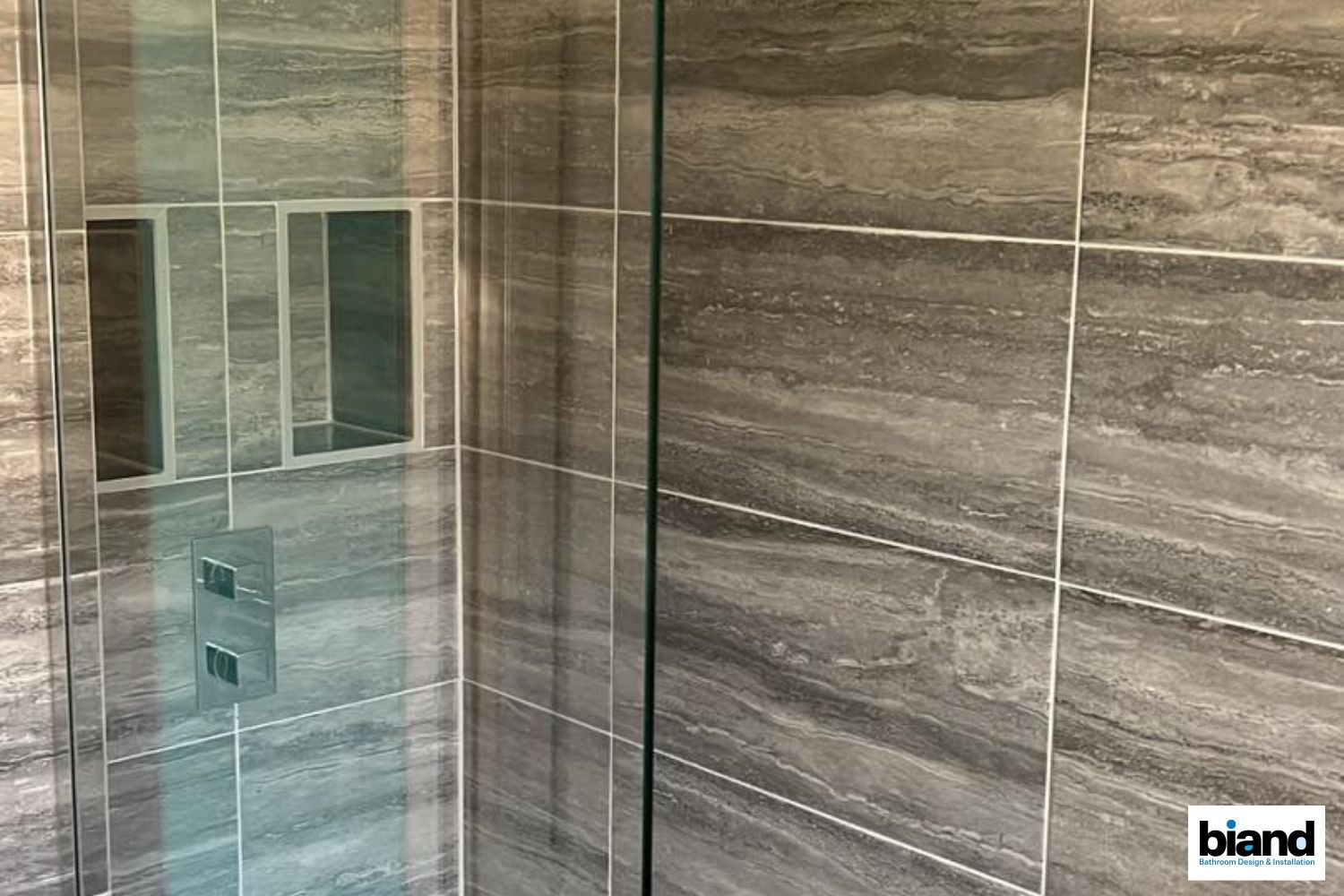 Dark gray tiled shower with glass door, recessed shelf, and chrome fixtures.