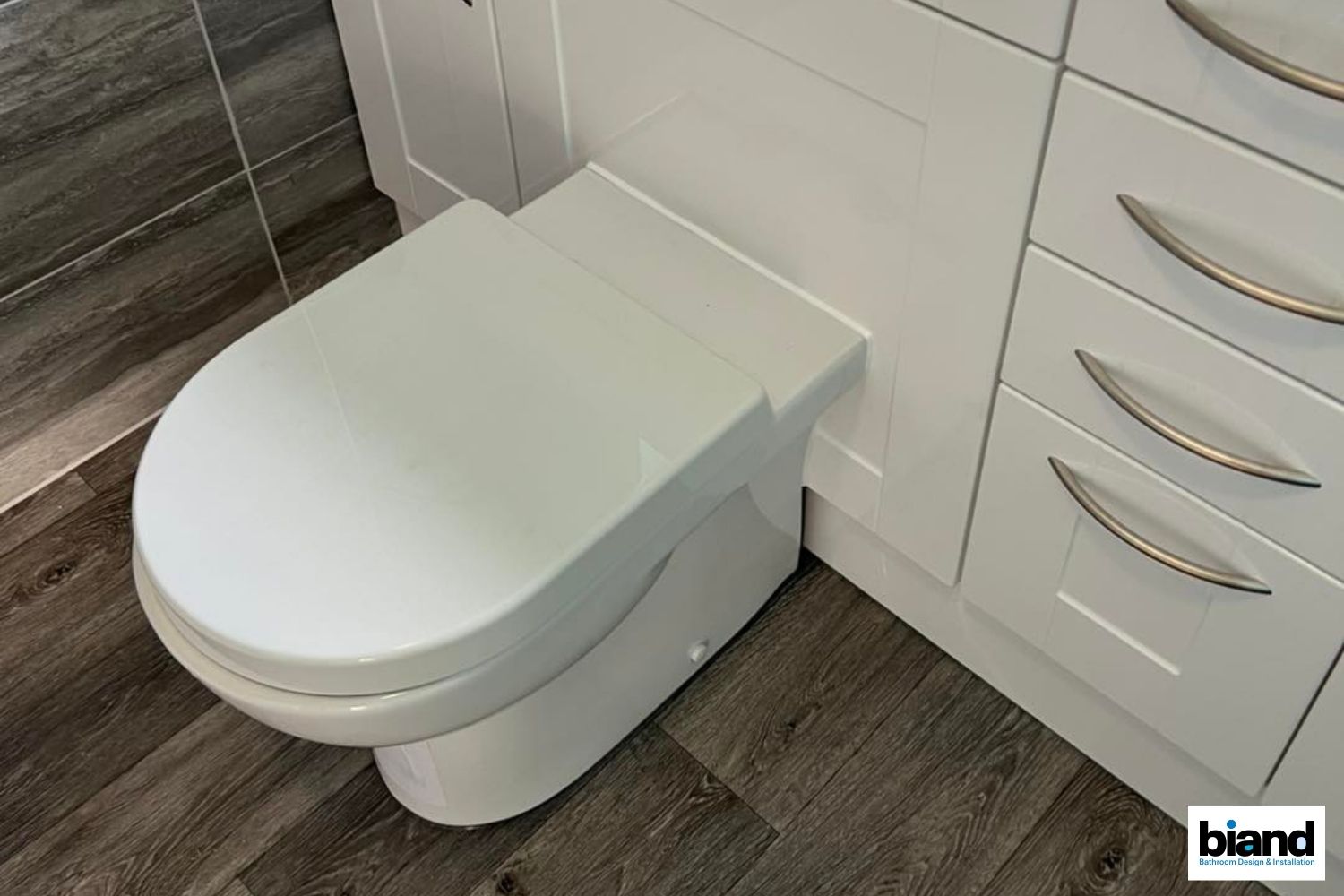 White toilet built into a white cabinet, next to drawers, on wood-look floor.