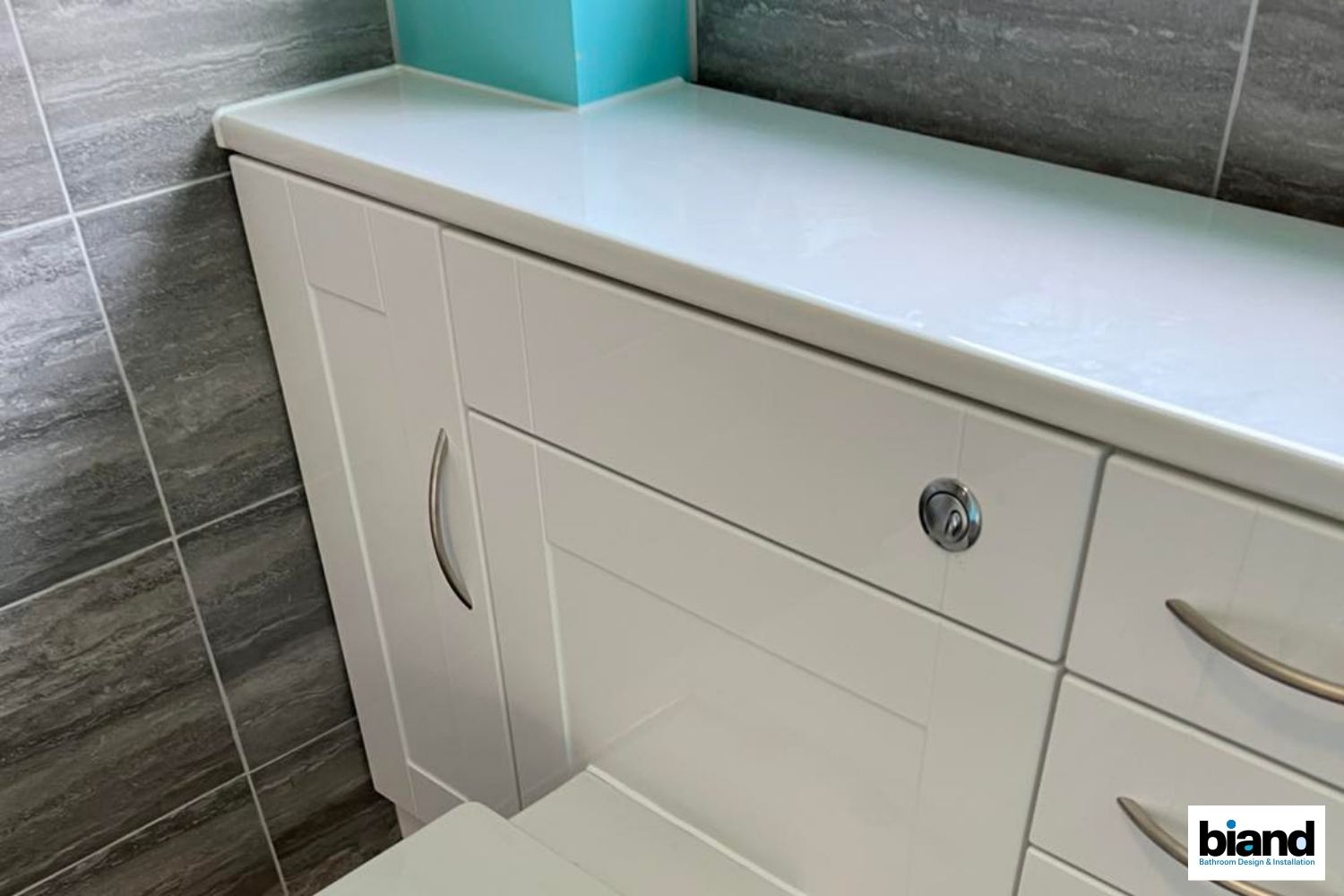 White bathroom cabinet with countertop and a teal box on top. Grey tiled wall.