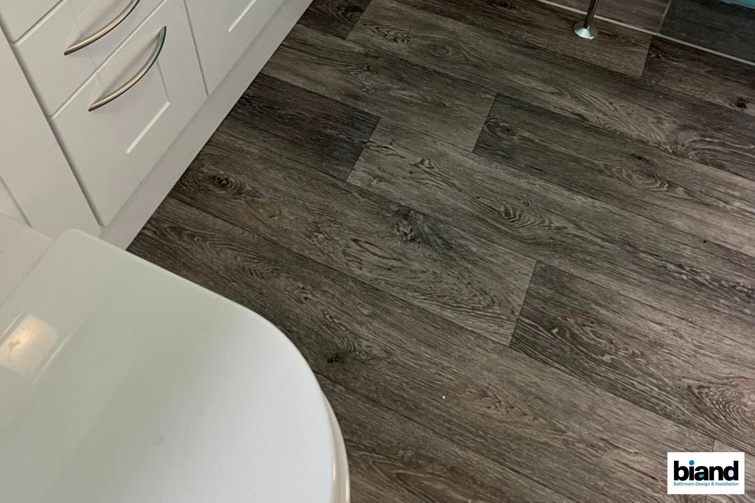 Bathroom floor with dark gray wood-look planks, near a white cabinet and toilet.