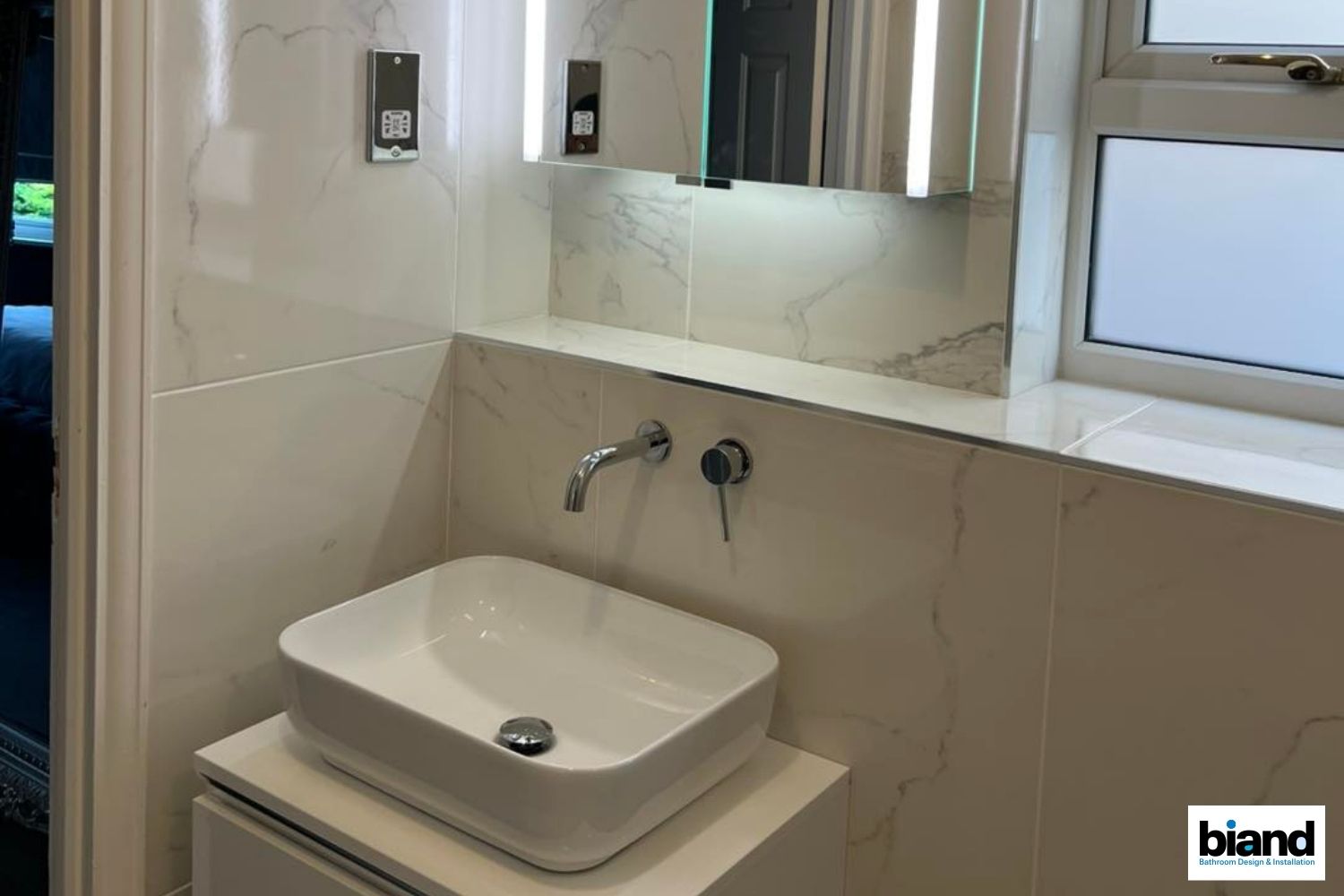 Modern bathroom with a white rectangular sink, marble-look tiles, and a mirror with lights above.