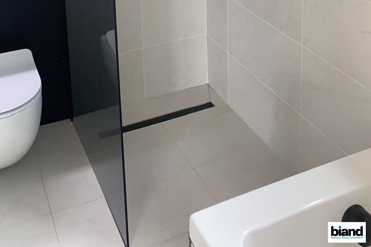 Bathroom with gray tiled floor, black drain, glass shower divider, toilet, and white sink.