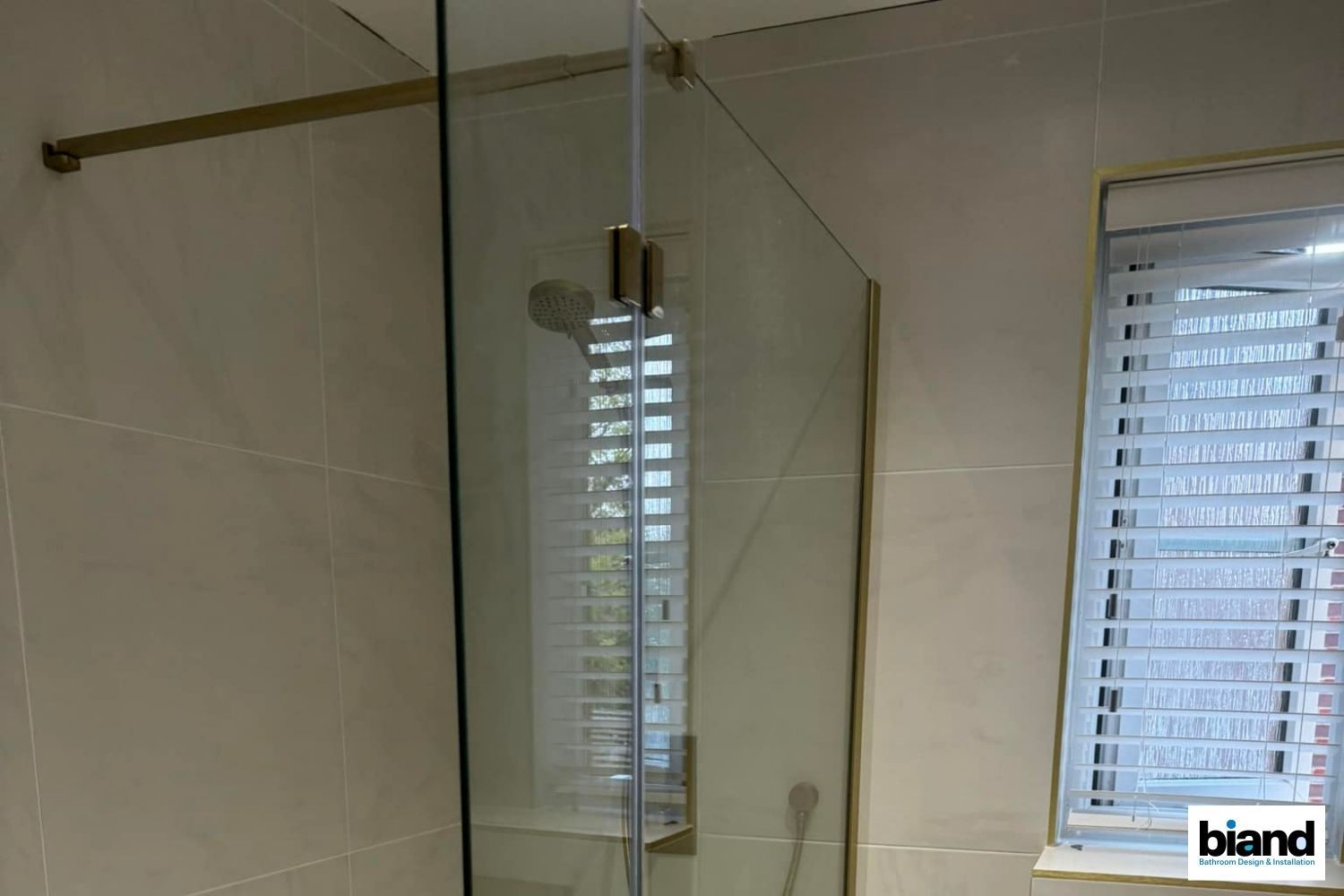 Shower enclosure with gold trim, a window with white blinds, and beige tiled walls.