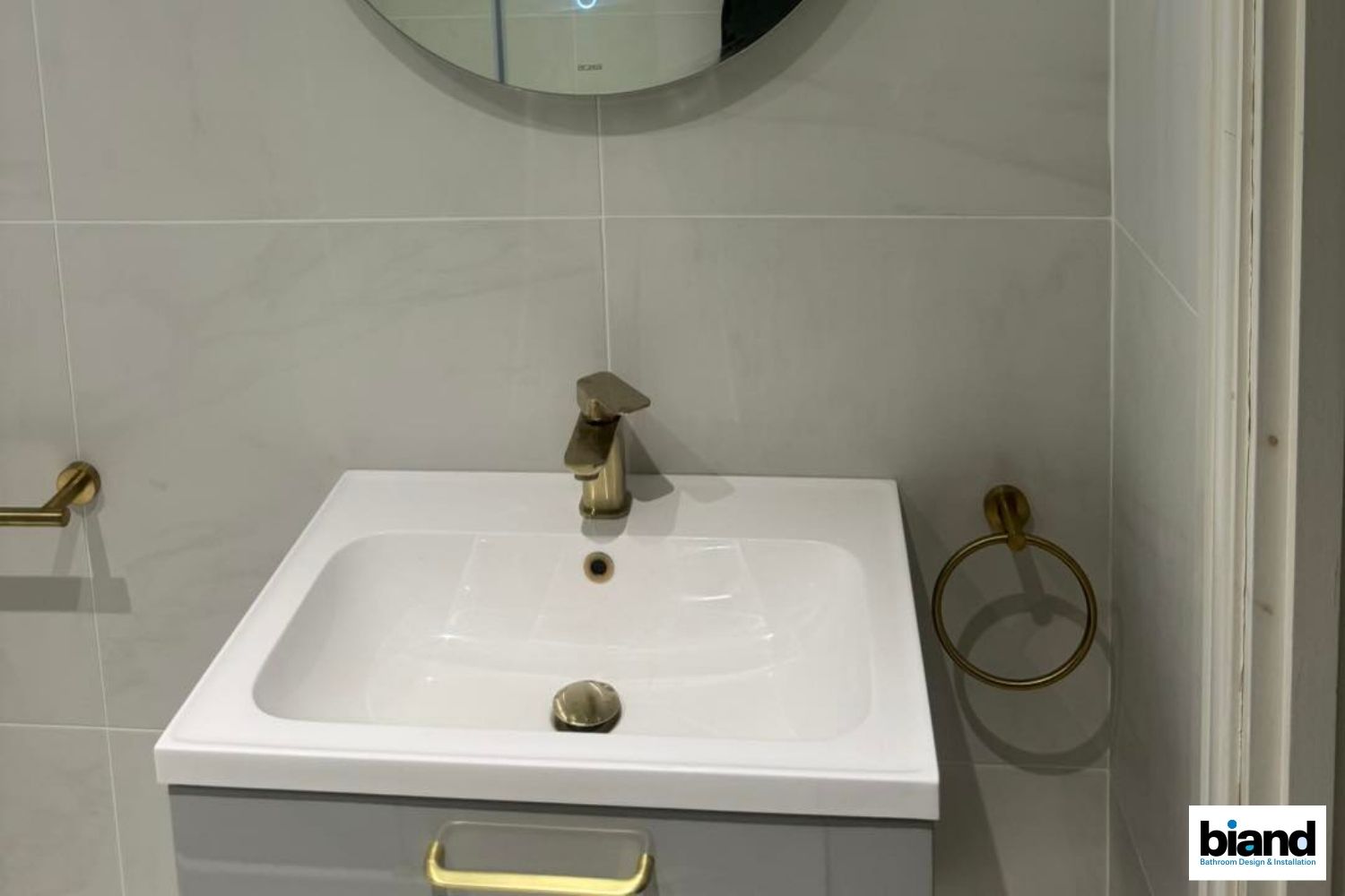 Bathroom with gold fixtures, grey vanity, white sink, and round mirror. Grey tile on the walls.