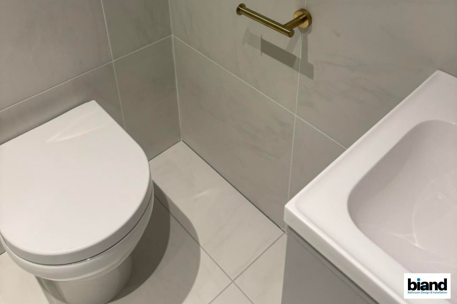 A small, modern bathroom with a white toilet, sink, and gold toilet paper holder. Light gray tiles on the walls and floor.
