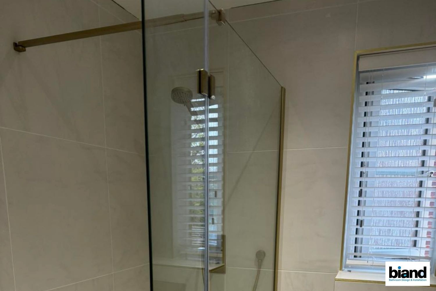 Gold-framed glass shower enclosure in a beige-tiled bathroom with a window and blinds visible in the background.