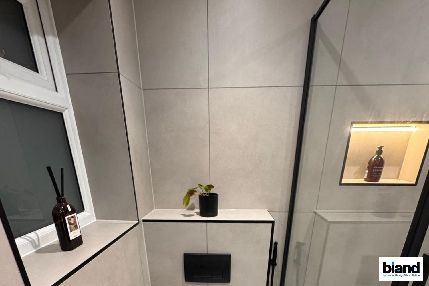 Bathroom interior with light gray tiles, black accents, small plant, and recessed shelf with bottle.