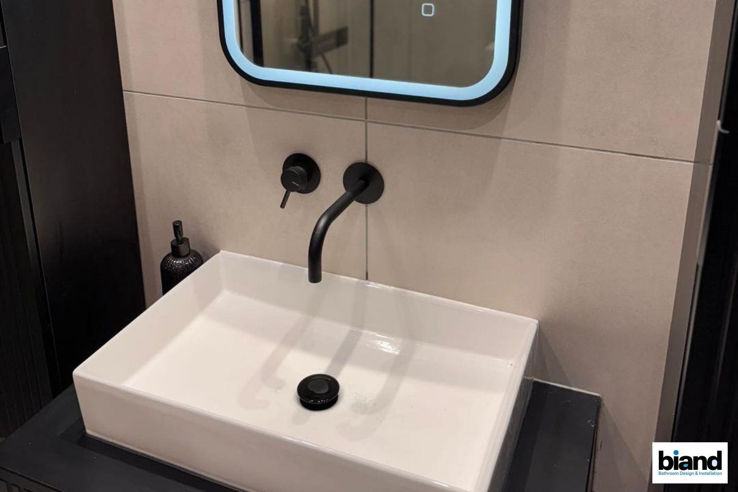 Black faucet and sink in a modern bathroom with a lighted mirror and neutral tiles.