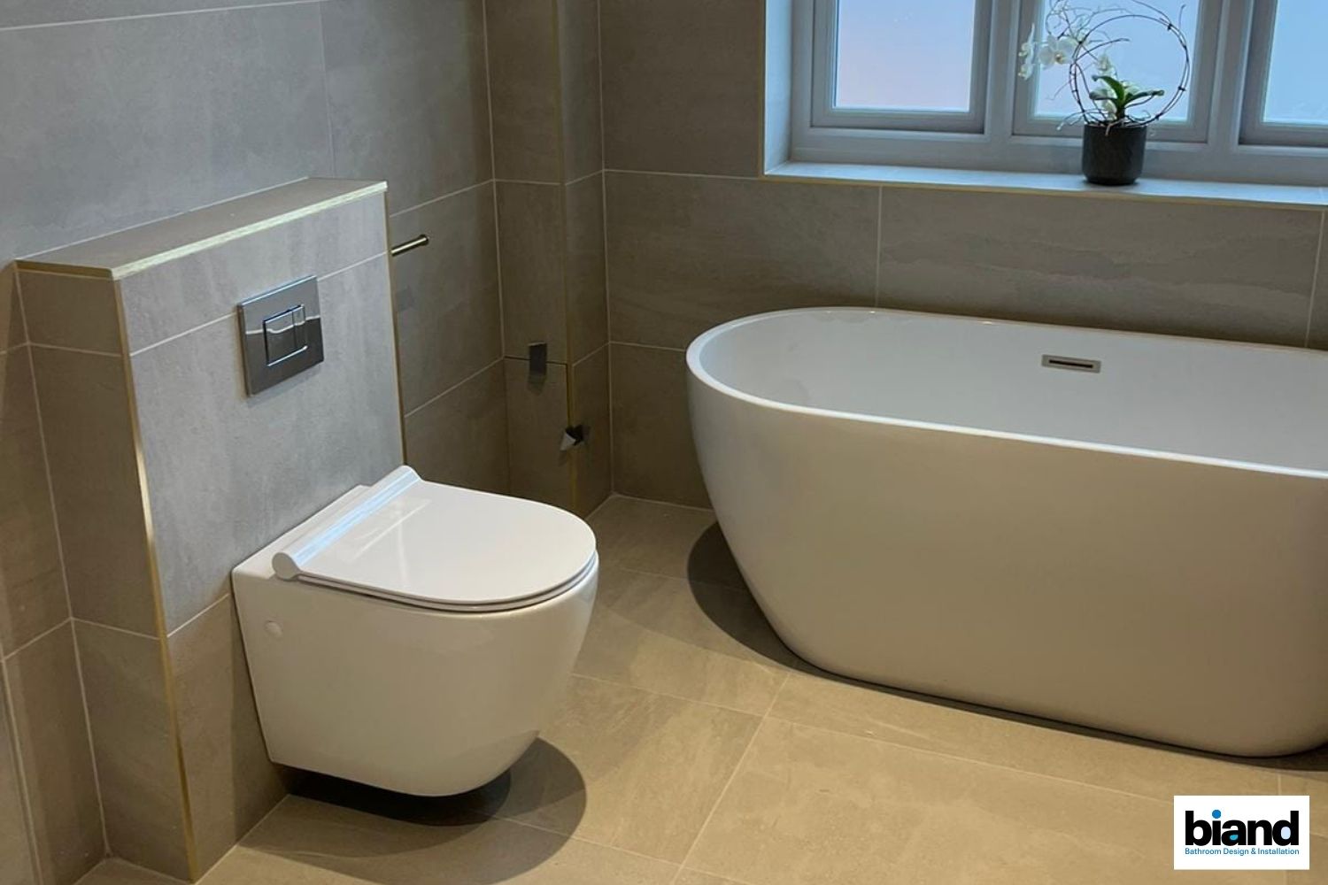 Modern bathroom with a wall-hung toilet, freestanding white bathtub, and a window. Neutral color scheme.