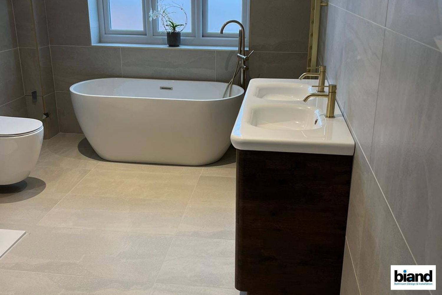 Modern bathroom with white tub, double sink on dark wood cabinet, and neutral tiles.