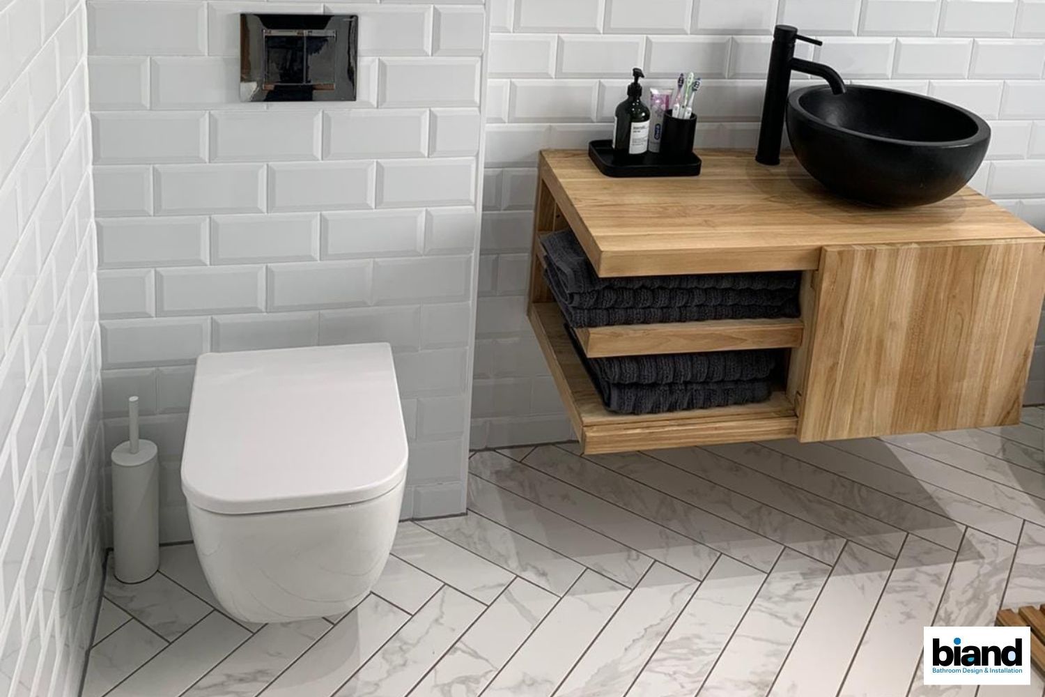 Modern bathroom: white subway tile walls, floating wood vanity with black bowl sink, gray towels, and white toilet.