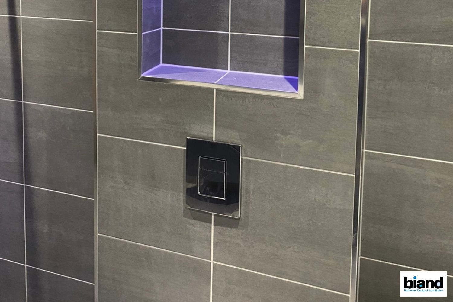 Gray tiled wall with a recessed shelf and black flush plate.