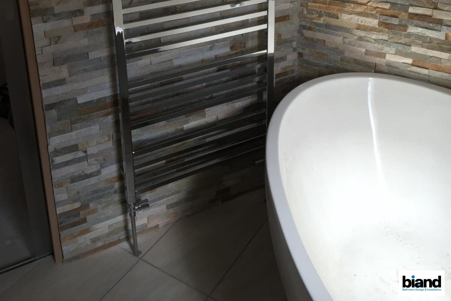 Chrome towel warmer on textured stone wall next to a white bathtub in a bathroom.
