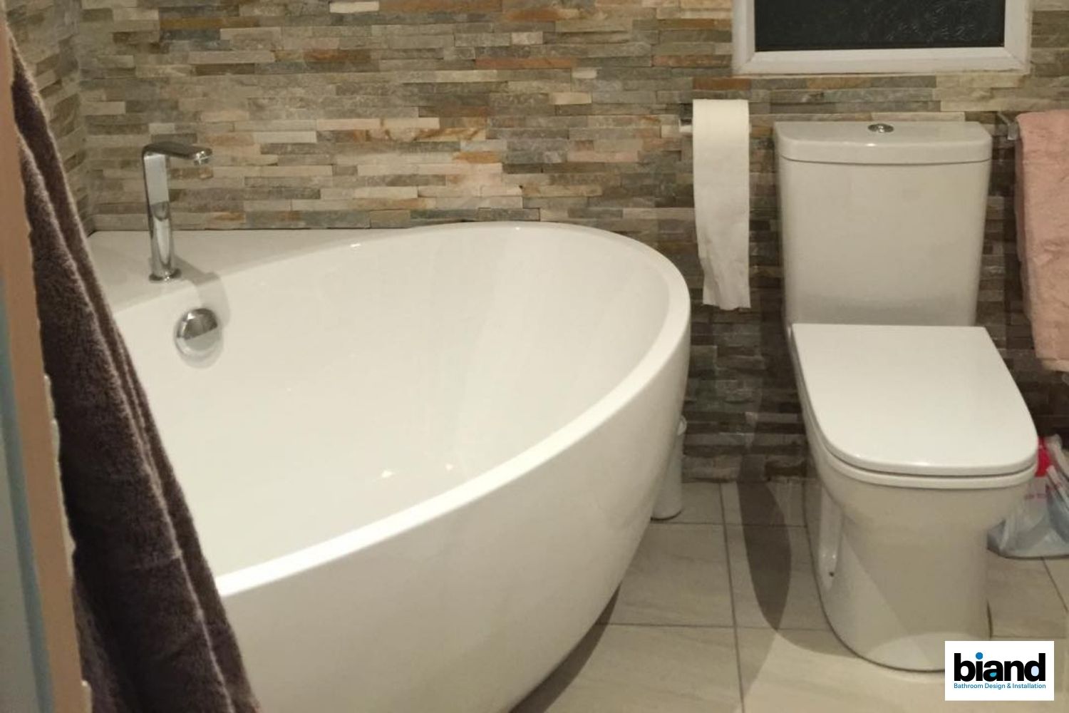 White bathtub and toilet in a bathroom with textured stone wall, towel, and toilet paper roll.