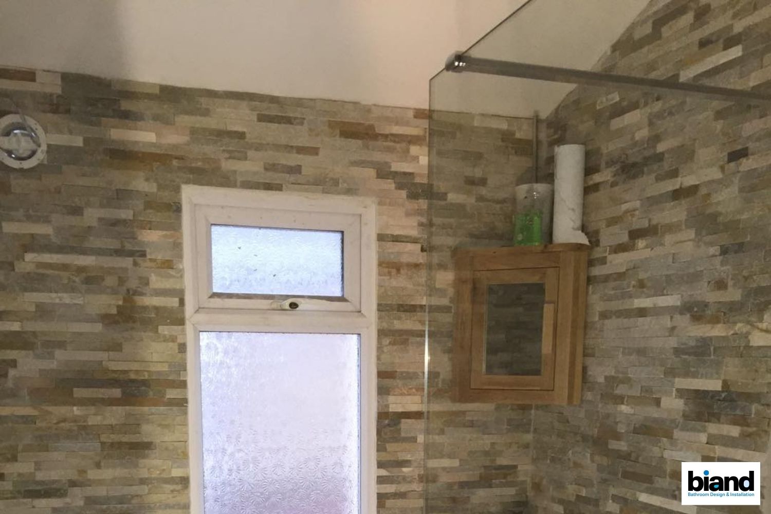 Bathroom with brick-style tiles, window, shower head and glass screen, and wooden cabinet.