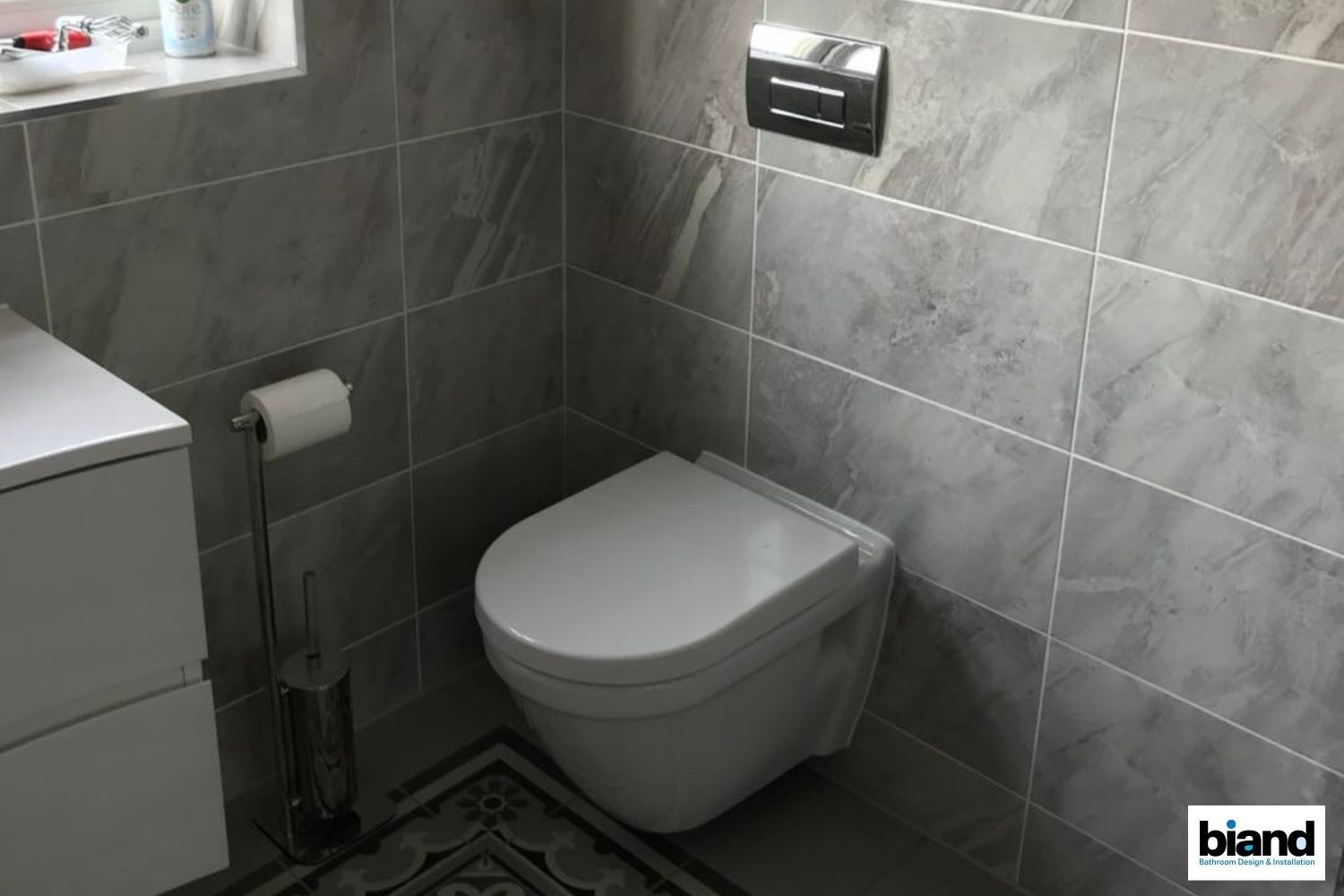 Gray-tiled bathroom with a wall-mounted toilet, flush panel, and toilet paper stand.