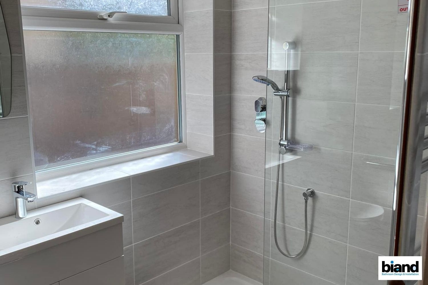 Bathroom with gray tiles, shower, vanity, and window with frosted glass.