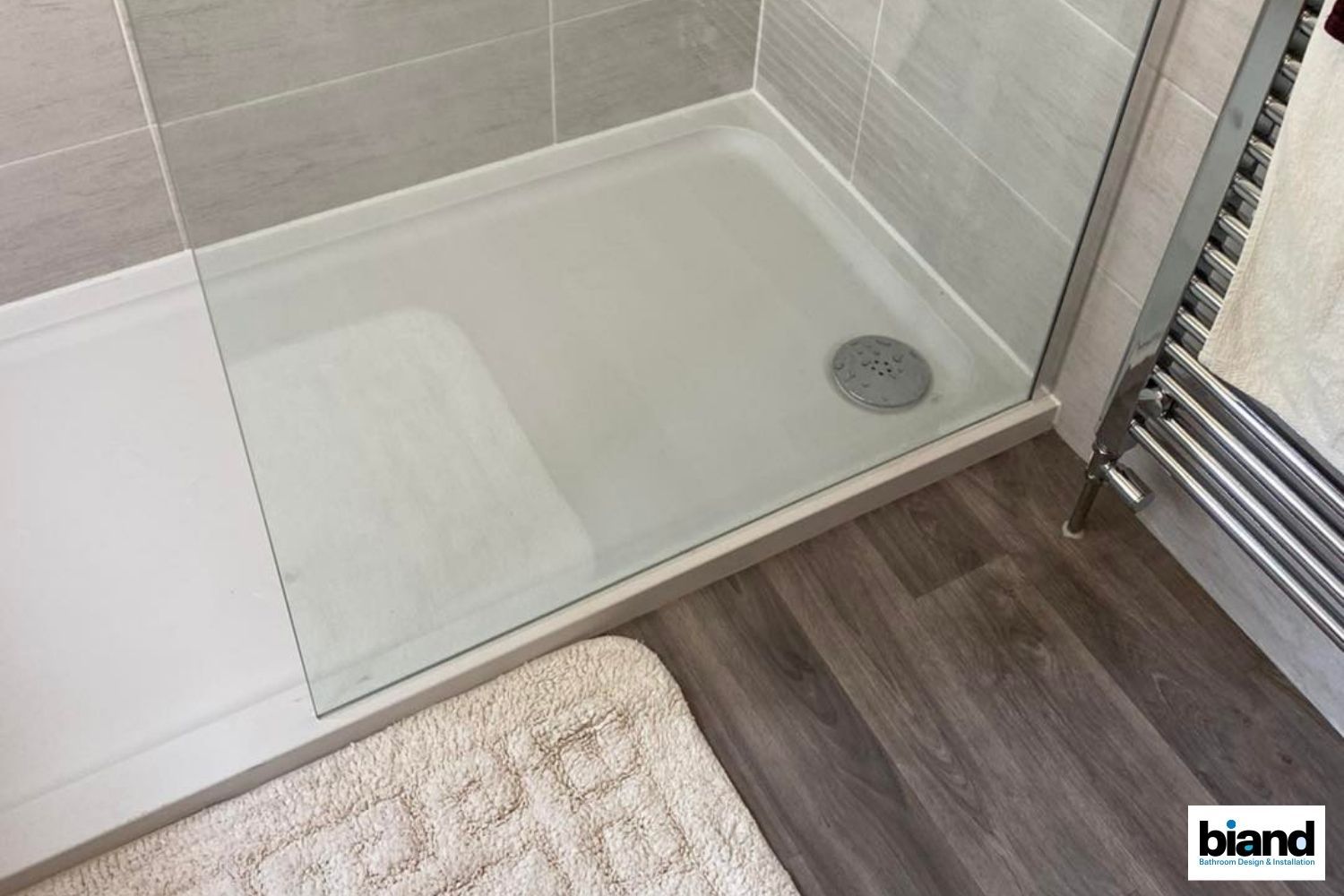 A shower with a glass door, white base, and gray tile walls. Wooden floor and a towel rack are visible.