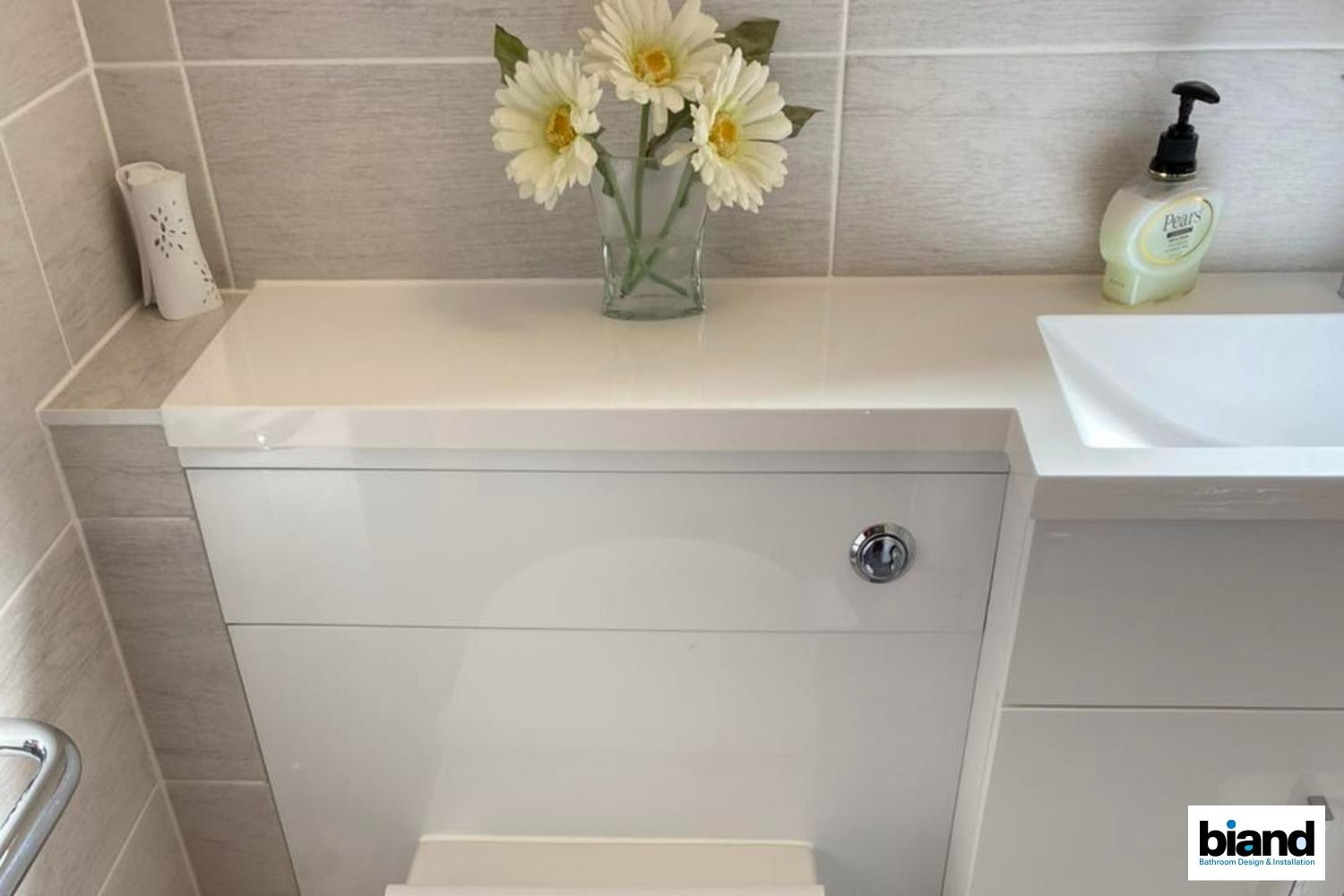 White, modern toilet with countertop holding flowers and soap, set in a tiled bathroom.