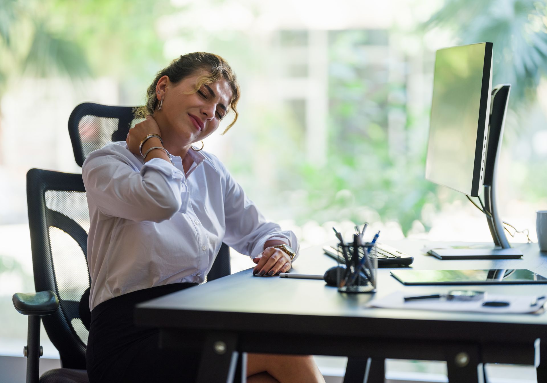 A woman is sitting at a desk in front of a computer holding her neck.