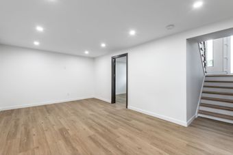 Empty modern room with light wood floors, white walls, recessed lights, and stairs at the right.