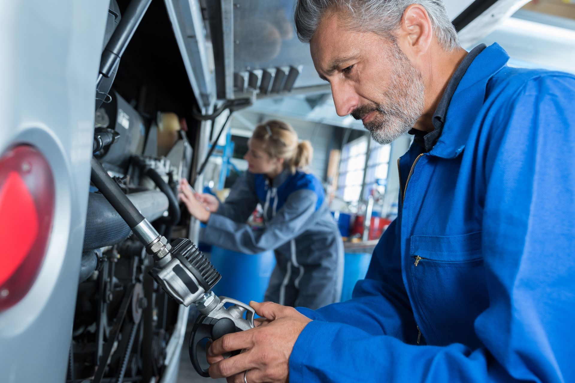 Mechanic in blue coveralls checking equipment inside a vehicle bay, with another worker blurred in the background