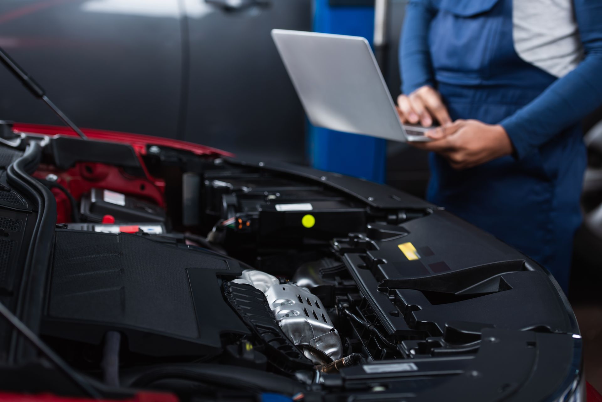 Open car hood with exposed engine while a person uses a laptop beside it