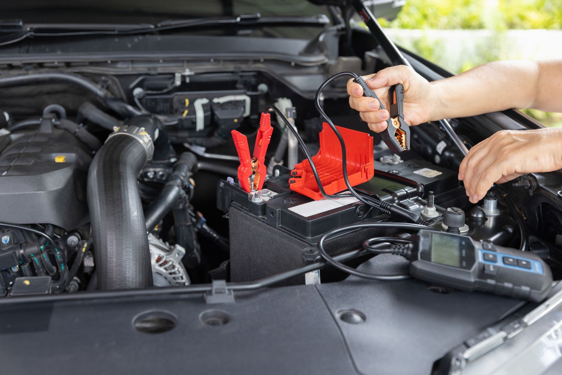 Car battery being jump-started in an engine bay with red cables and a hand holding a jumper clamp