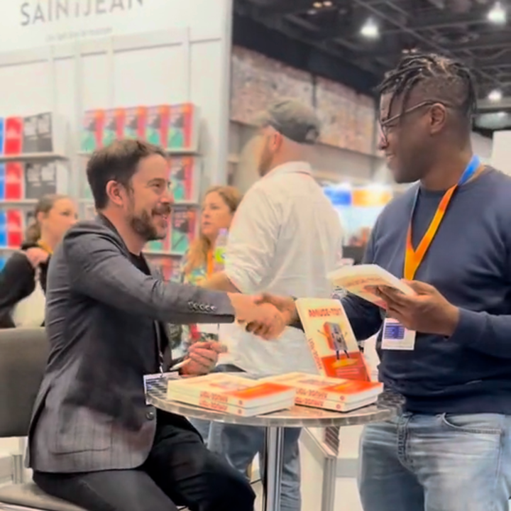 Deux hommes se serrent la main devant un stand de librairie, avec des livres empilés et un décor de salon professionnel.