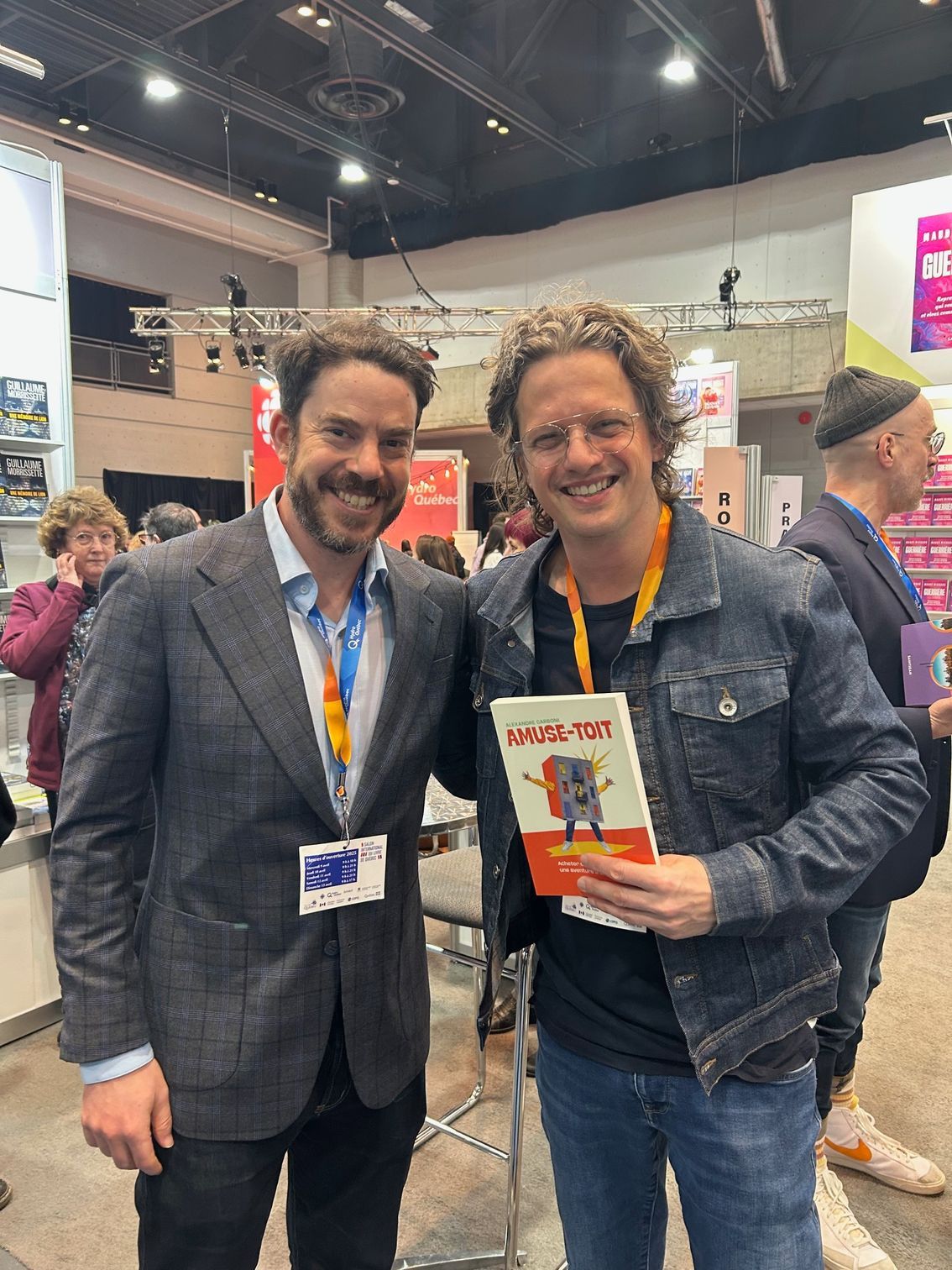 Deux hommes, souriants et tenant un livre, se trouvent dans un hall d'exposition bondé lors d'un congrès.