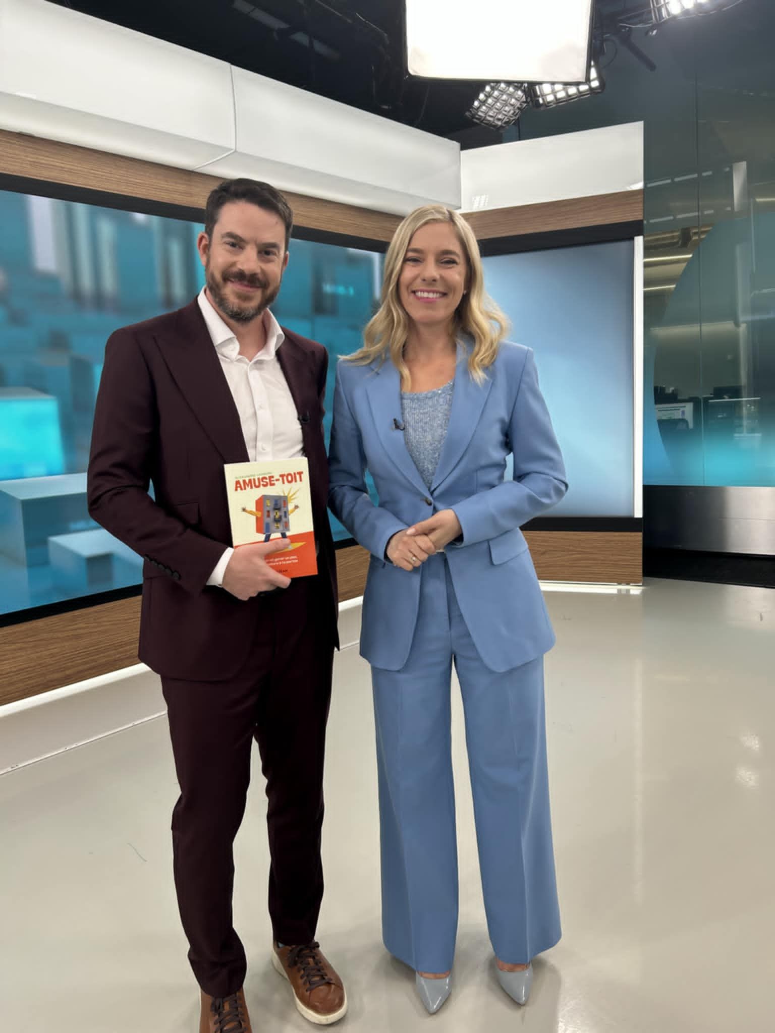 Deux présentateurs se tiennent dans un studio, l'un tenant un livre, devant des écrans bleus et sous des lumières vives.