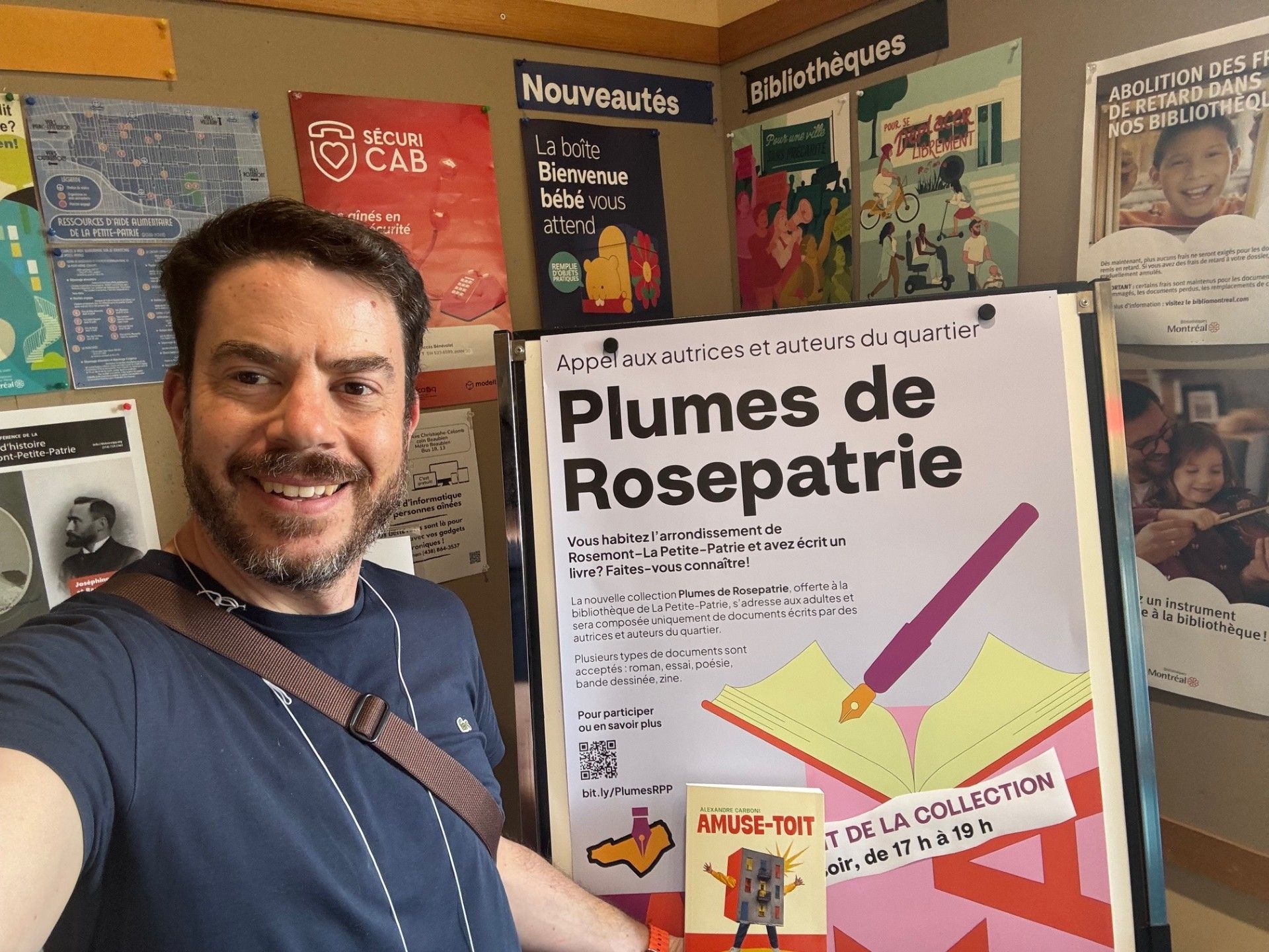 Un homme sourit à côté d'une affiche intitulée « Plumes de Rosepatrie » dans une salle de classe ou une bibliothèque colorée.
