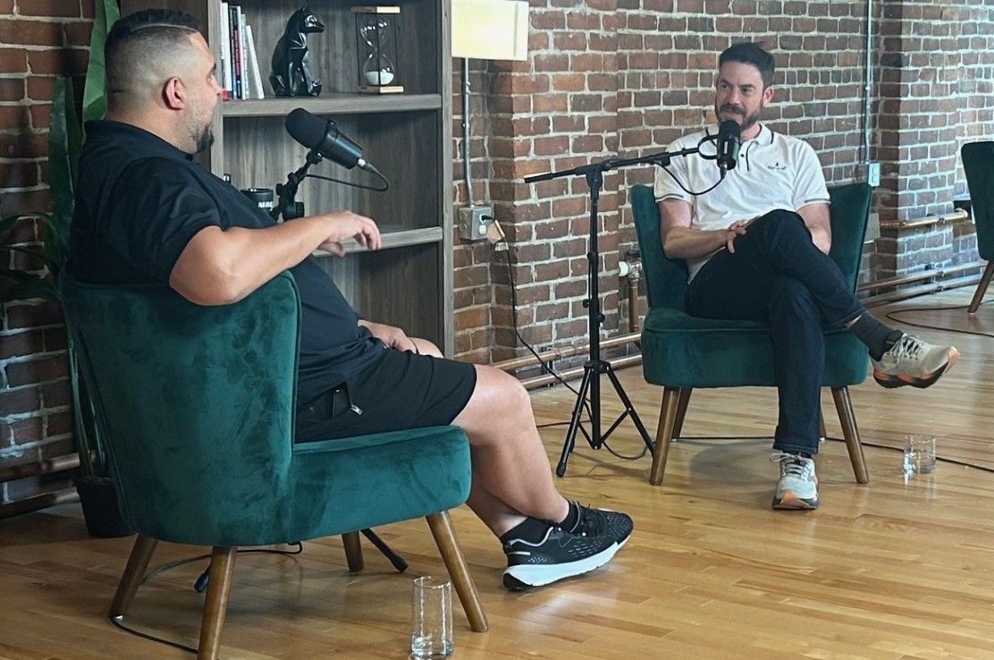 Deux hommes assis sur des chaises vertes enregistrent une interview pour un podcast dans un studio aux murs de briques.