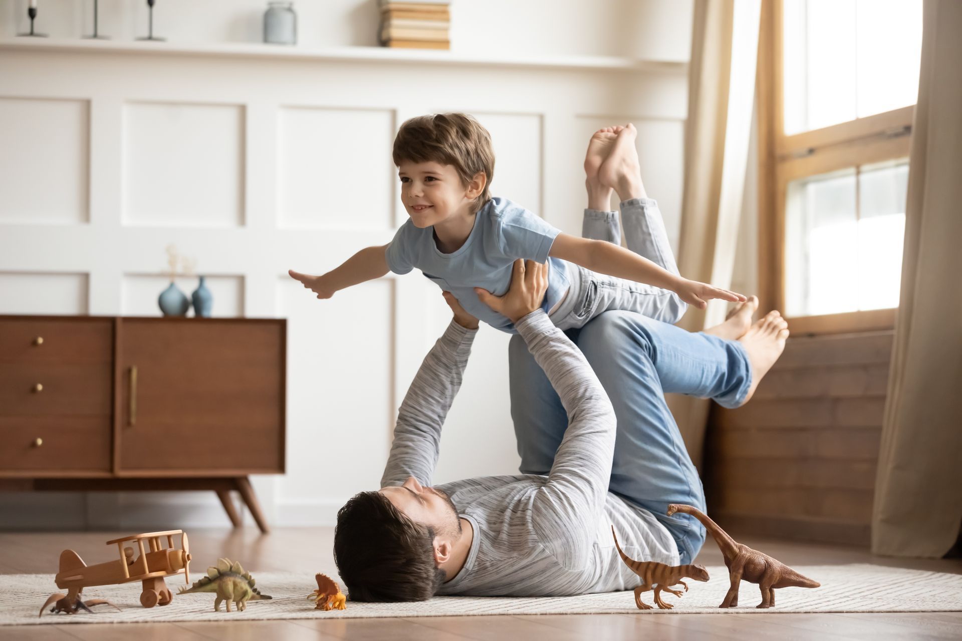 Joyful young man father lying on carpet floor, lifting excited happy little child son at home
