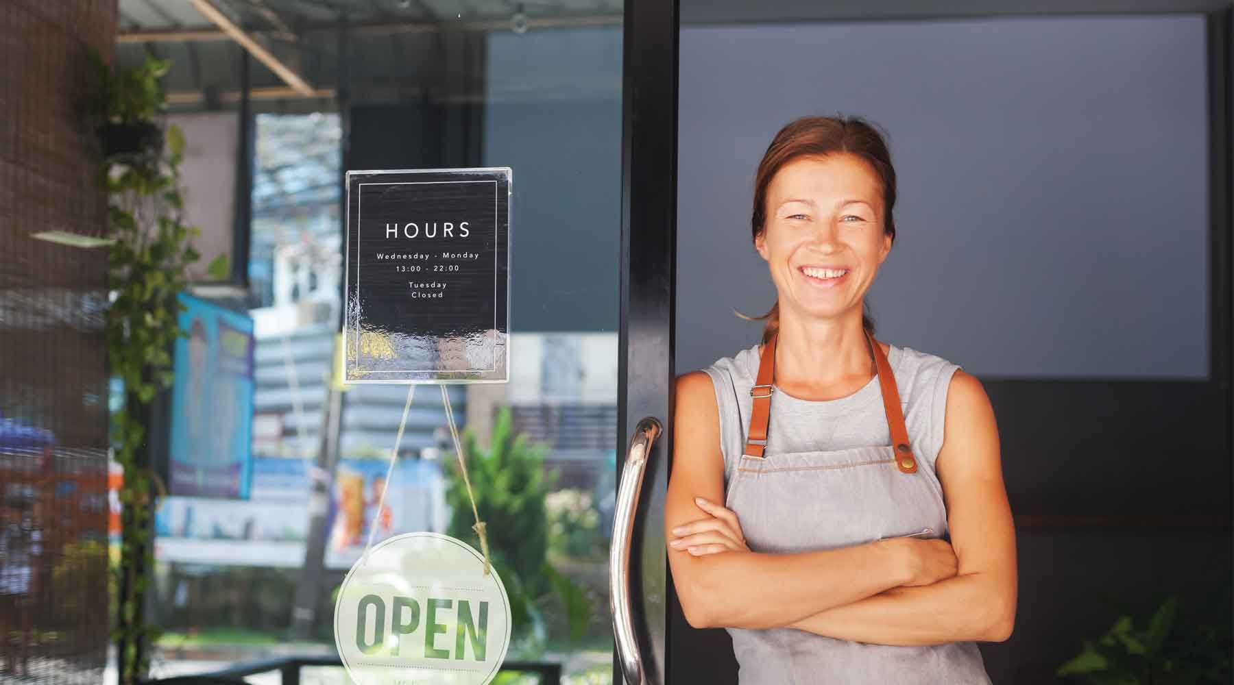 Woman in apron smiles, arms crossed, standing in doorway of a shop with