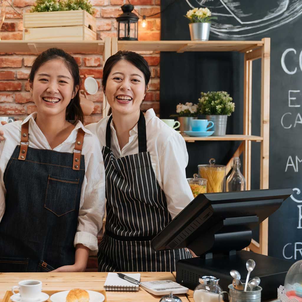 Two women in aprons smiling behind a cafe counter.