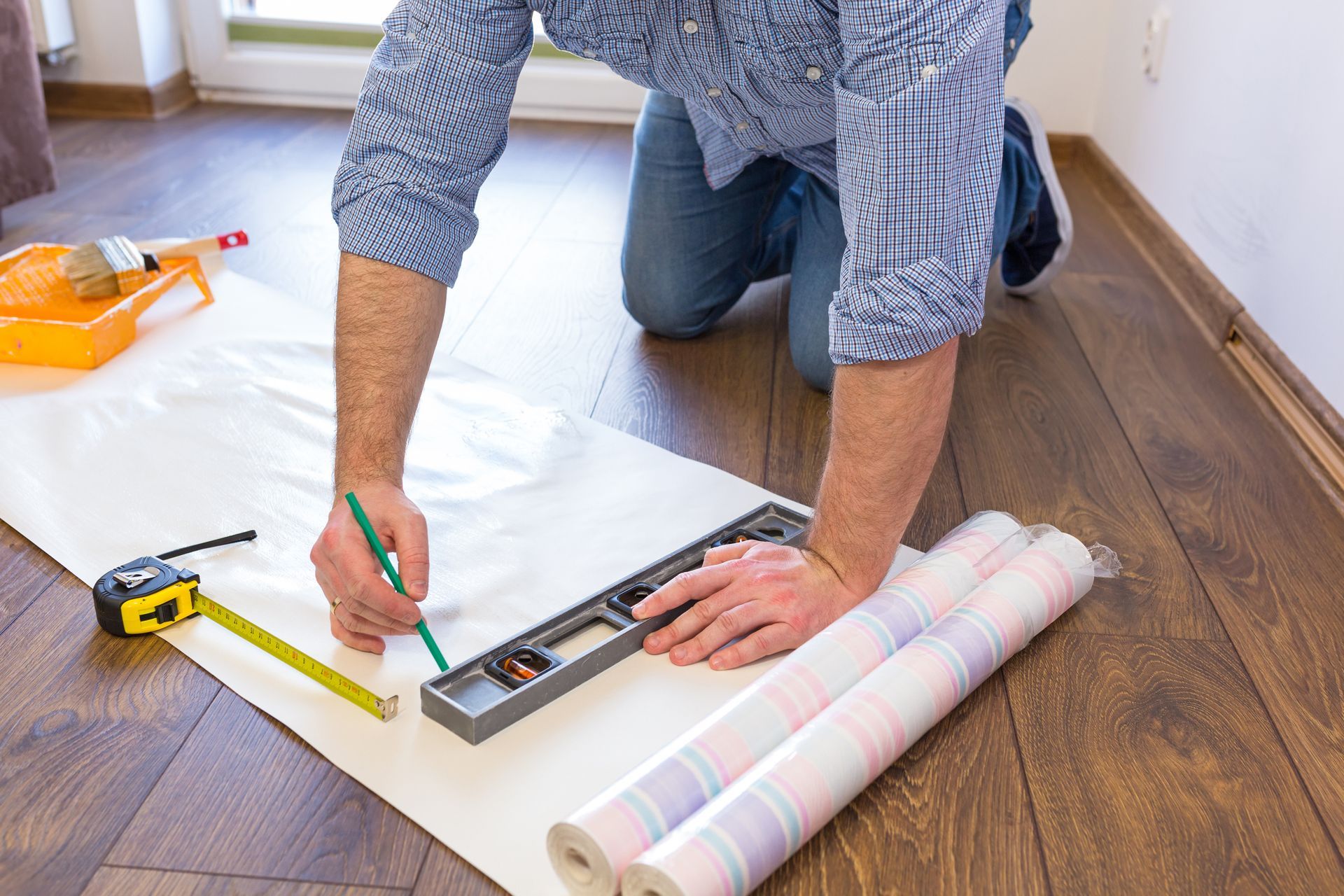 Person kneeling on wood floor measuring wallpaper with a ruler and pencil beside a tape measure.
