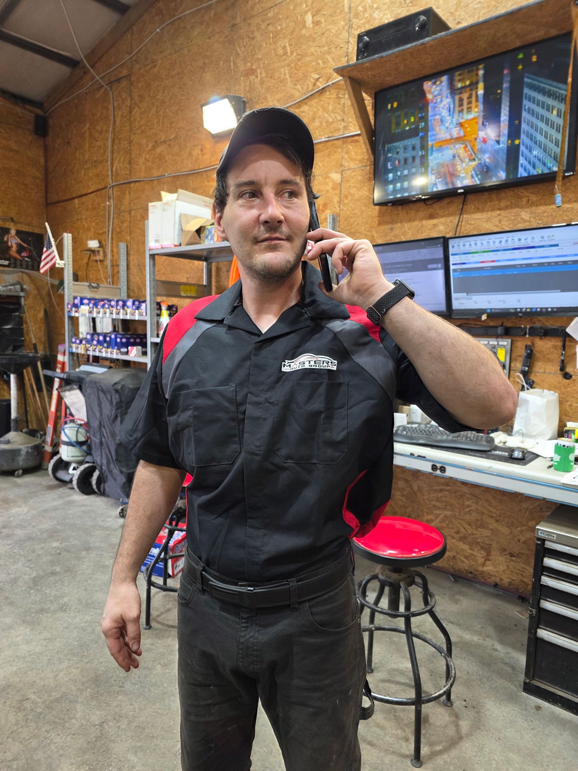 Person in a black shirt and cap talks on a phone in a workshop with monitors and tools. | Masters Auto Group