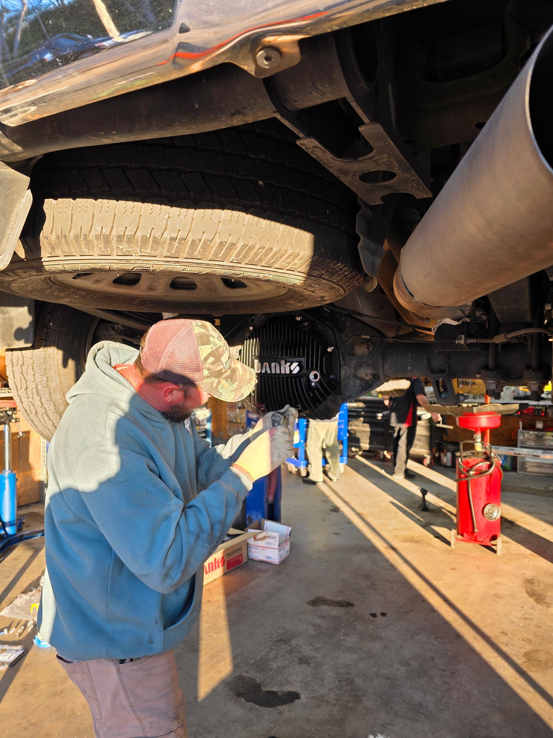Person in a blue hoodie working under a vehicle in a garage, with a spare tire and exhaust pipe visible. | Masters Auto Group