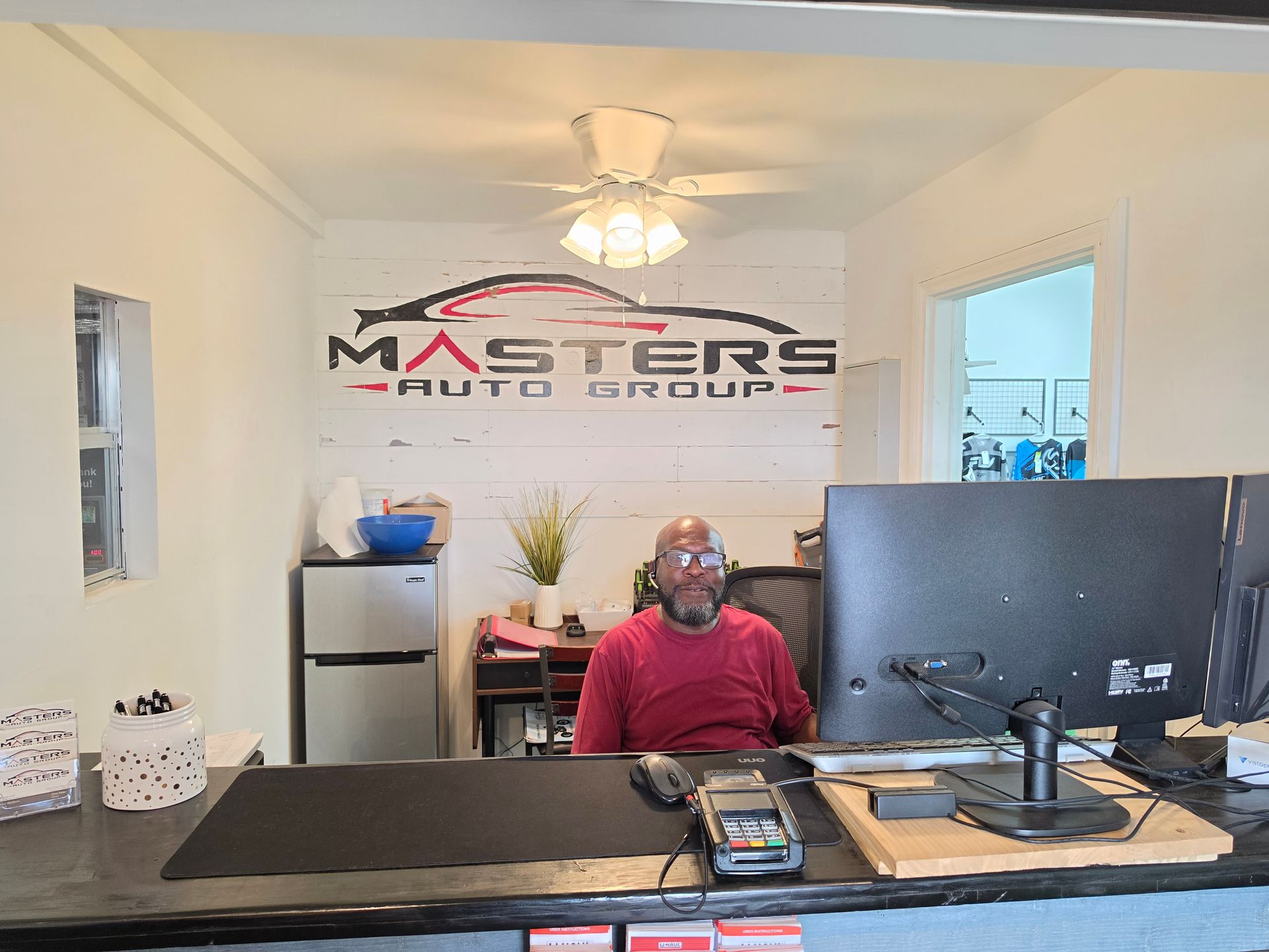 Person seated at a desk in a bright office with a Masters logo on the wall and a large monitor in front | Masters Auto Group