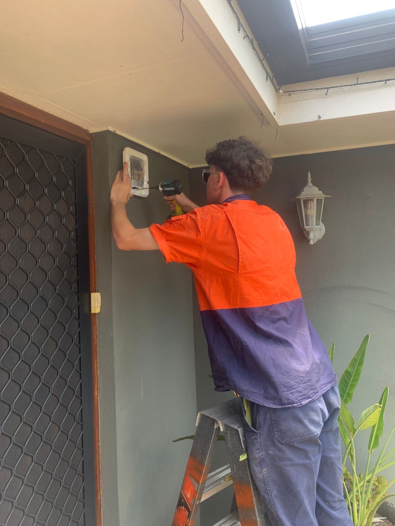 Person in Orange/blue Work Shirt Using a Drill on a Wall, Standing on a Ladder Near a Door — Frontier Security in Gatton, QLD
