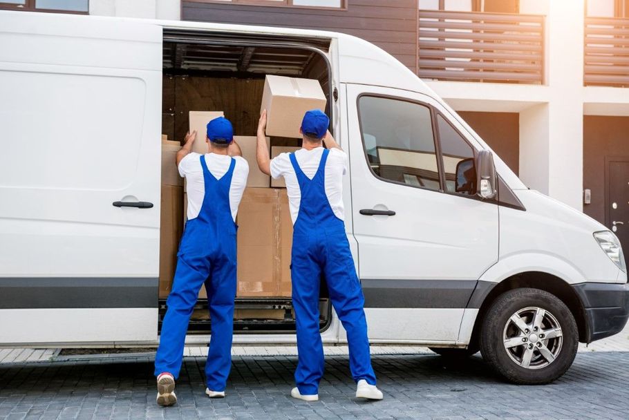 Two movers in blue overalls unload boxes from a white delivery van