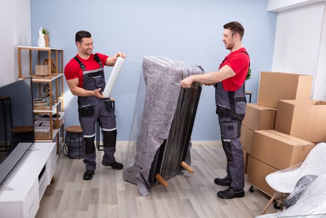 Two movers in red shirts carrying a wrapped mattress through a room with boxes and shelves