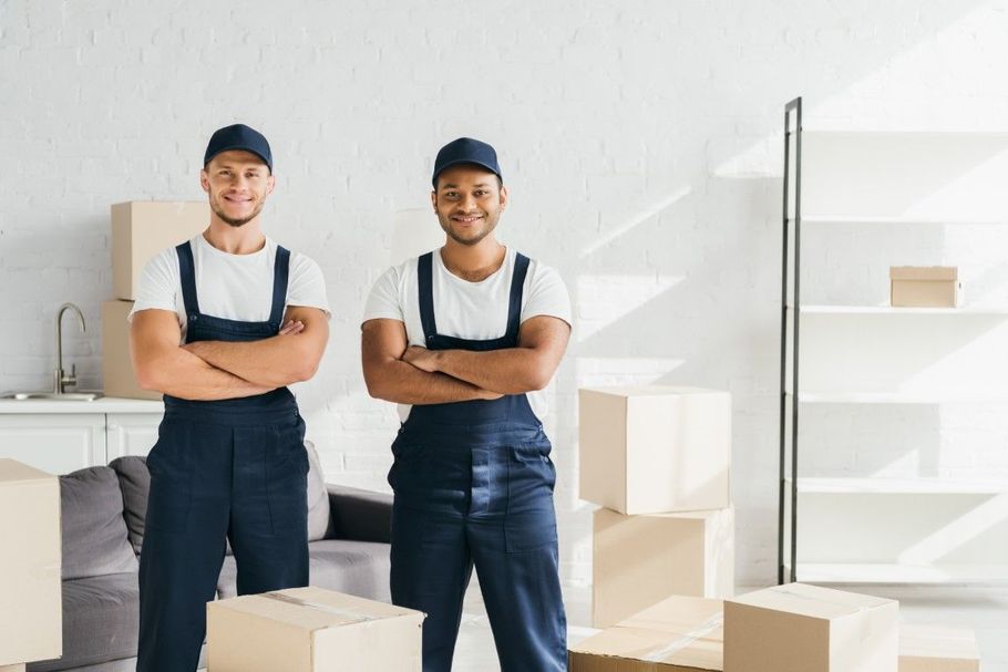 Two movers in navy overalls stand with arms crossed in a bright room with stacked moving boxes.