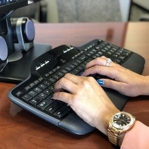 Hands typing on a black ergonomic keyboard, gold watch on wrist.