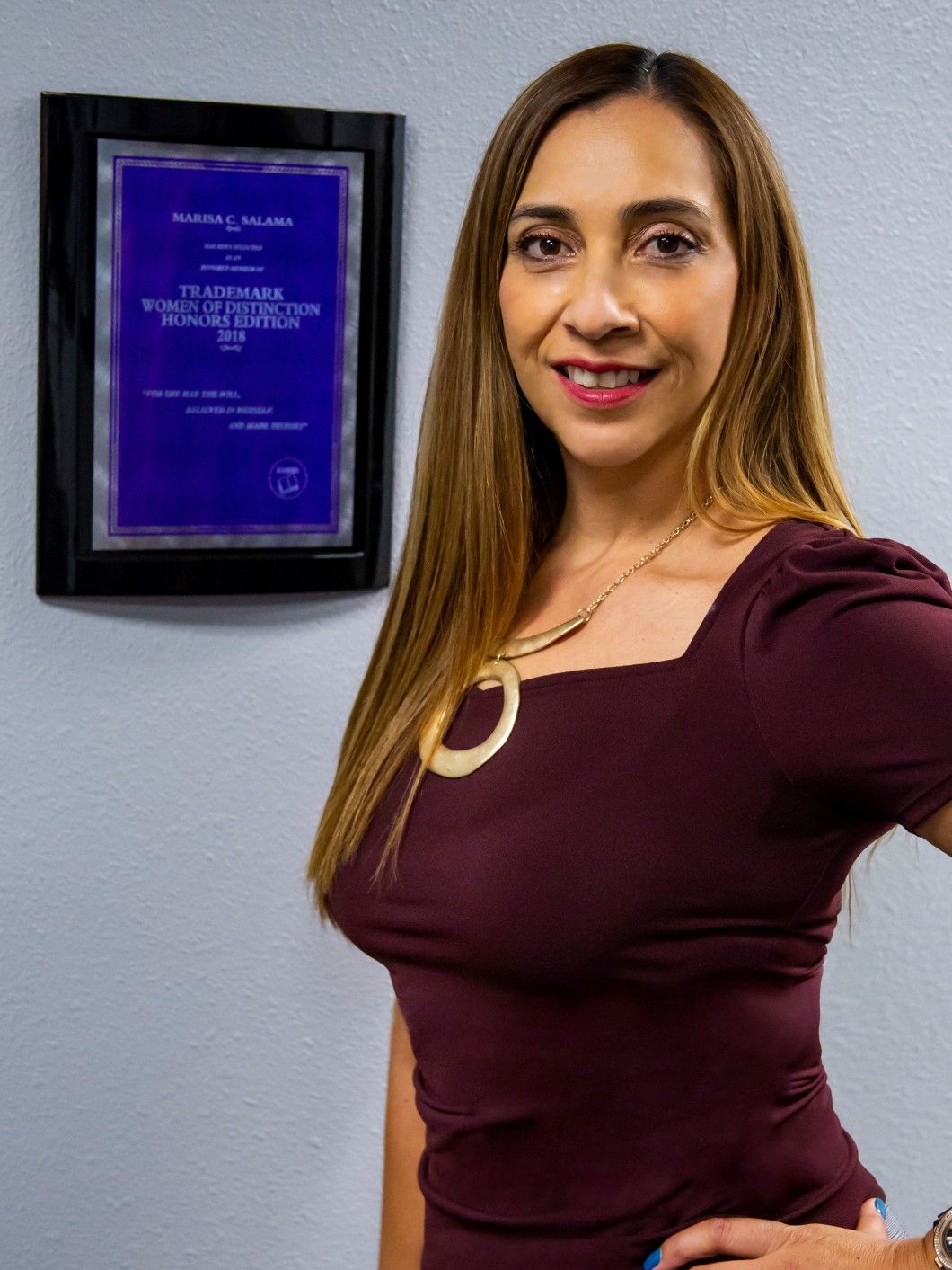 Woman with long brown hair, wearing a burgundy top and gold necklace, stands near a framed certificate.