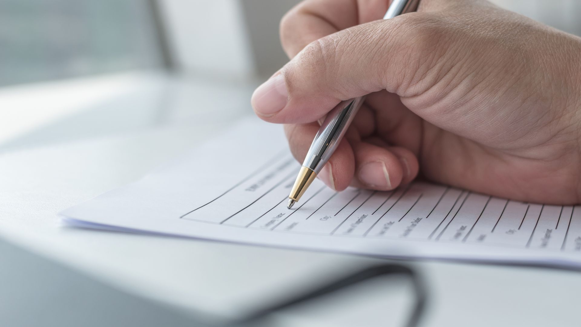 Hand writing on a form with a pen over a blurred desk background