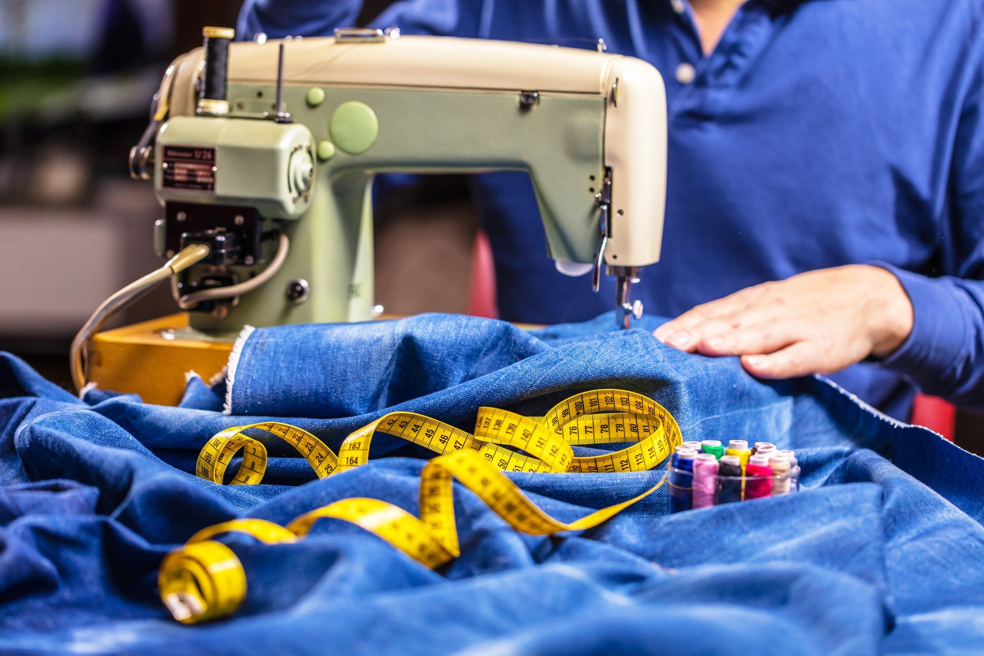 Vintage sewing machine with blue fabric, yellow measuring tape, and a person in a blue shirt sewing