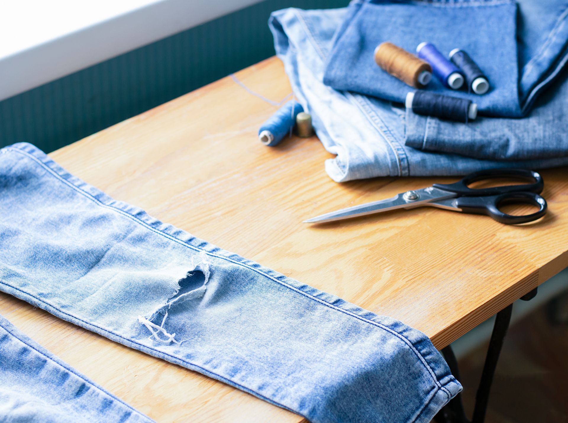 Fabric and sewing tools on a wooden table, with jeans and blue denim laid out for alterations.